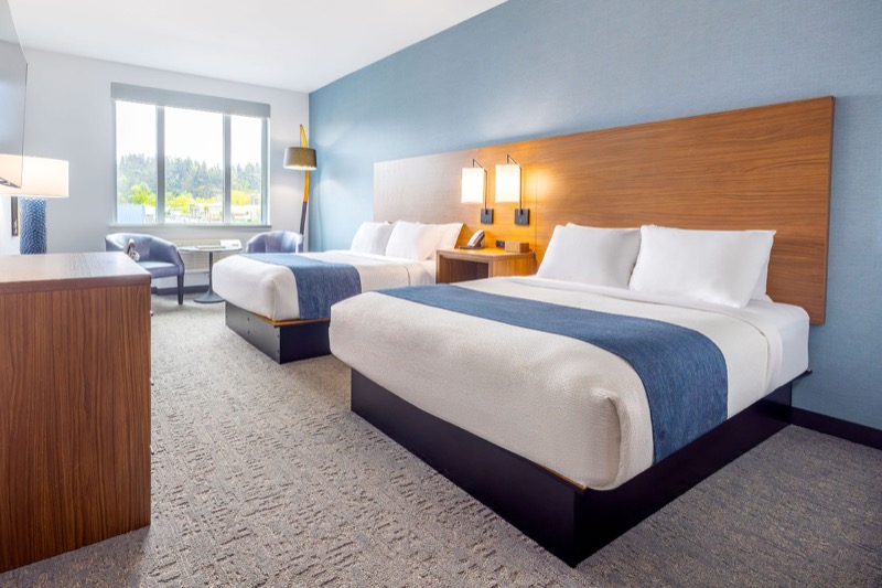 ADA Double Queen guest room with two beds, blue runners, warm wood headboard with sconces, seating area, and large window with treetop view