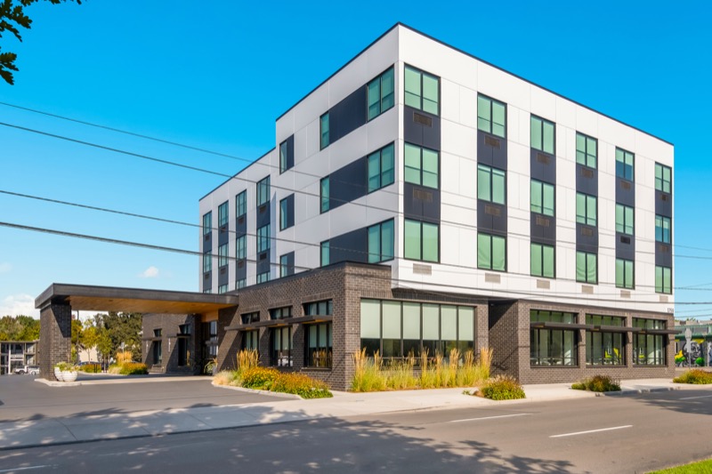 Exterior of the modern multi-story hotel, white and dark facade, brick base, and covered entrance under a clear blue sky