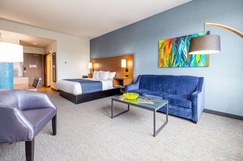Deluxe Suite living area with blue velvet sofa, glass coffee table, bold colorful abstract art, arc floor lamp, and king bed in the background