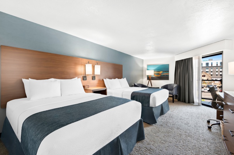 Double Queen guest room with two beds, teal accent wall, warm wood headboard with sconces, armchair, desk, and large window with city view