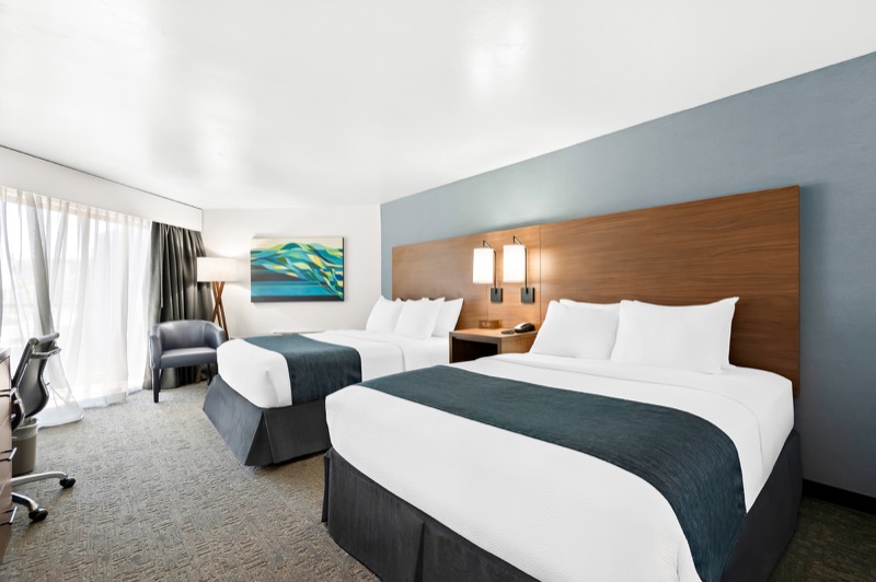 Double Queen guest room from the opposite angle, two beds with navy runners, wood headboard, seating corner, and large window with sheer curtains