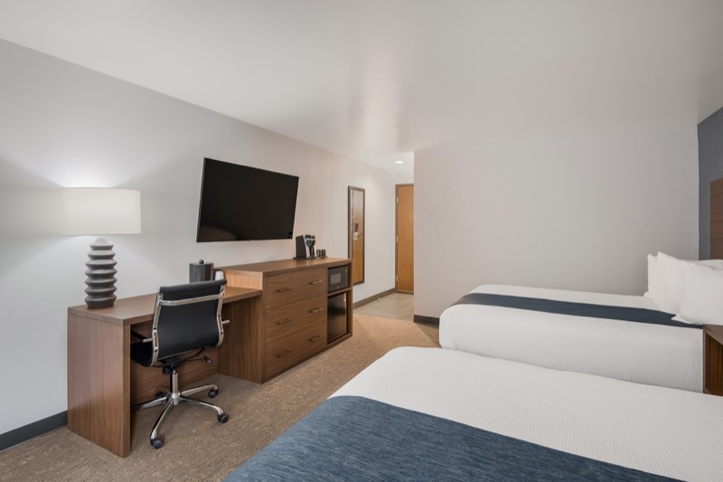 Double Queen room with two beds, wooden desk with office chair, dresser, wall-mounted TV, and hallway leading to entrance door