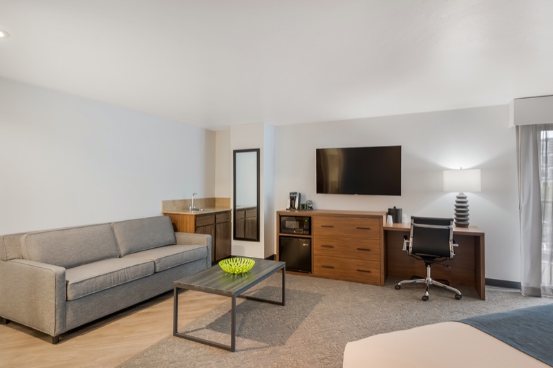 King Suite living area with grey sofa, glass coffee table, wall-mounted TV, dresser with mini-fridge, desk, and kitchenette nook