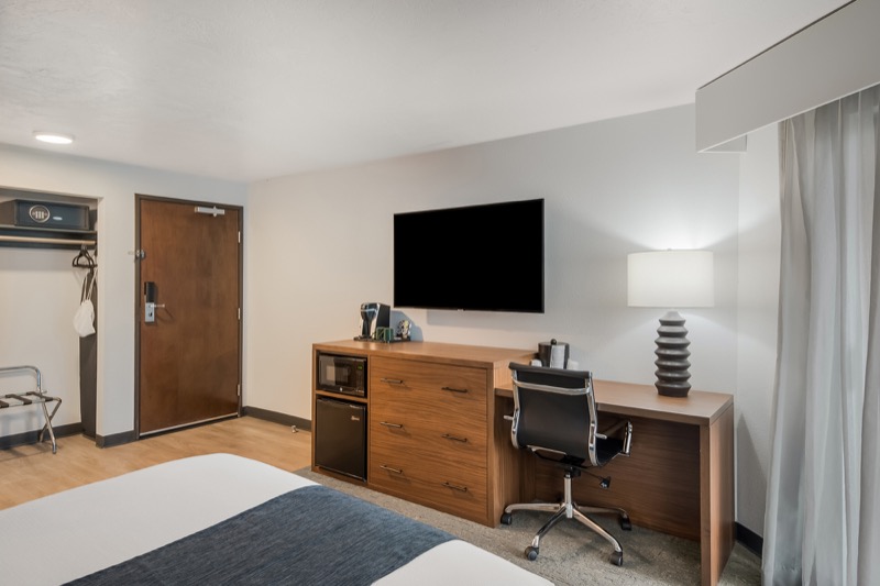 Queen guest room with wall-mounted TV, wood dresser and desk, black office chair, microwave, mini-fridge, and corner of the queen bed
