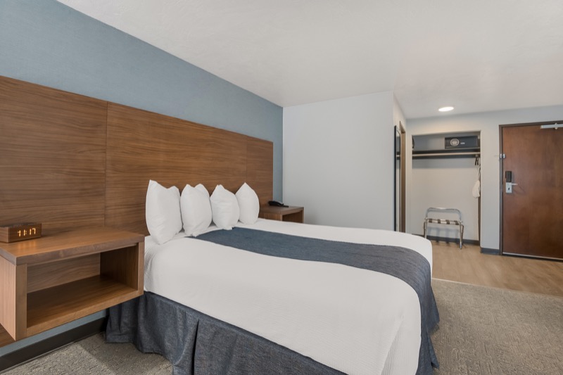 Queen guest room with queen bed, light blue accent wall, wide wood headboard, floating nightstands, alarm clock, and closet entryway