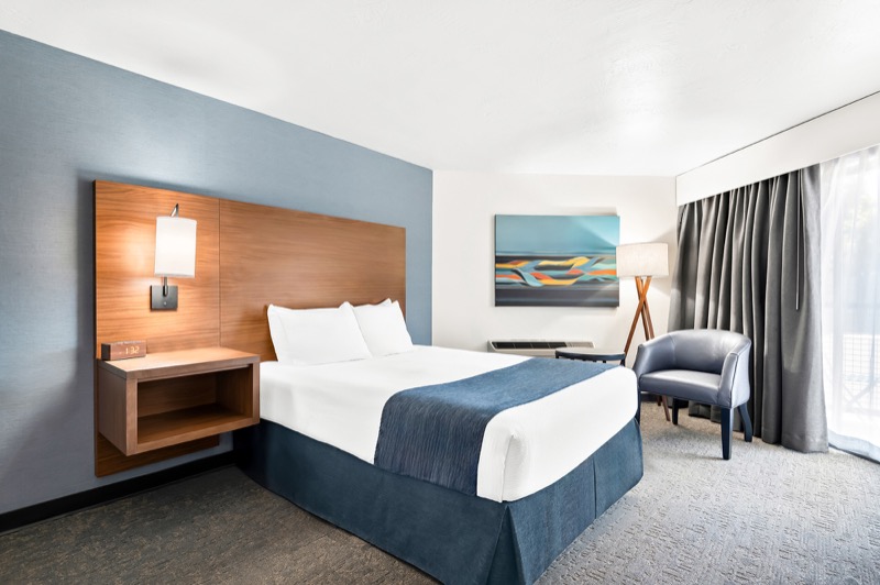 Queen guest room with queen bed, blue accent wall, wood headboard, abstract art, and blue armchair by the large window