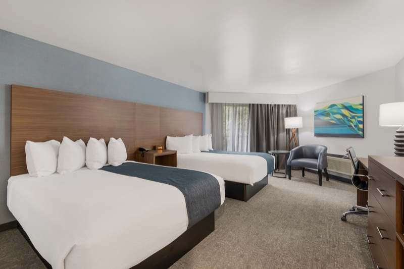 Waterview Double Queen guest room with two beds, blue-grey accent wall, wood headboard, seating area, abstract art, and desk with dresser