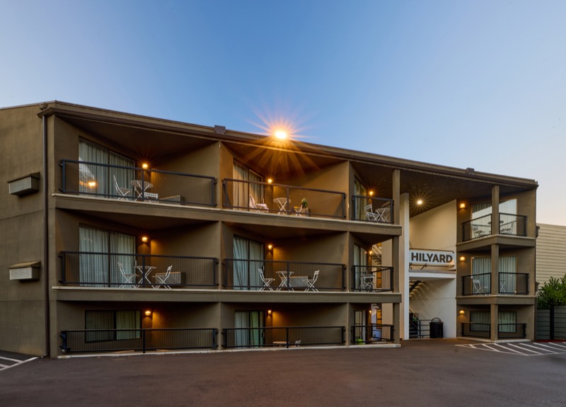 Exterior of the Hilyard hotel at dusk, three levels of balconies with warm lighting, black railings, and outdoor seating